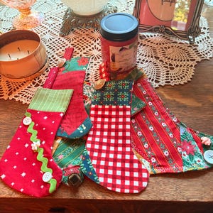 May include: A collection of four fabric Christmas stockings featuring various patterns including red with white stars, red and white gingham, and red with green floral stripes. Each stocking is embellished with decorative buttons and rickrack trim.