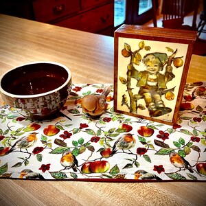 May include: A brown ceramic bowl, a small brown bird figurine, and a rectangular wooden box with a tile insert depicting a child sitting in an apple tree, all arranged on a white fabric placemat featuring a pattern of red apples and birds.