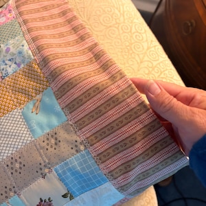 May include: A patchwork pillow cover featuring a variety of fabric squares in shades of blue, pink, yellow, and white with floral, plaid, and geometric patterns. The border is made of a tan fabric with a repeating dark brown floral stripe pattern.
