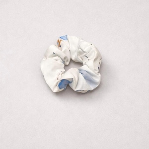 May include: White scrunchie with a blue and tan floral pattern. The fabric is gathered, creating a textured, circular shape. The scrunchie is a hair accessory, perfect for tying up hair.