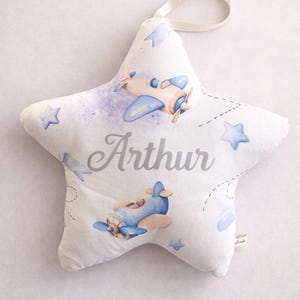 May include: A white star-shaped cushion with a ribbon loop. It features illustrations of blue and tan aeroplanes, blue stars, and the name "Arthur" in silver lettering. The design has a watercolour style.