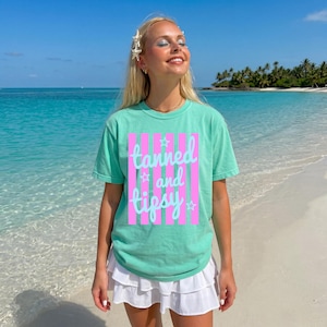 May include: A mint green t-shirt with the words "tanned and tipsy" in a pink and white striped design. The shirt is worn with a white skirt on a beach with turquoise water and a blue sky.