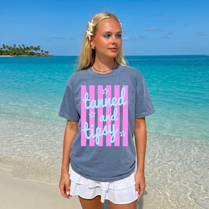 May include: A blue-grey t-shirt with the words "tanned and tipsy" in a light blue script, set against a pink striped background. The shirt is worn with a white skirt. The background shows a tropical beach with clear blue water.