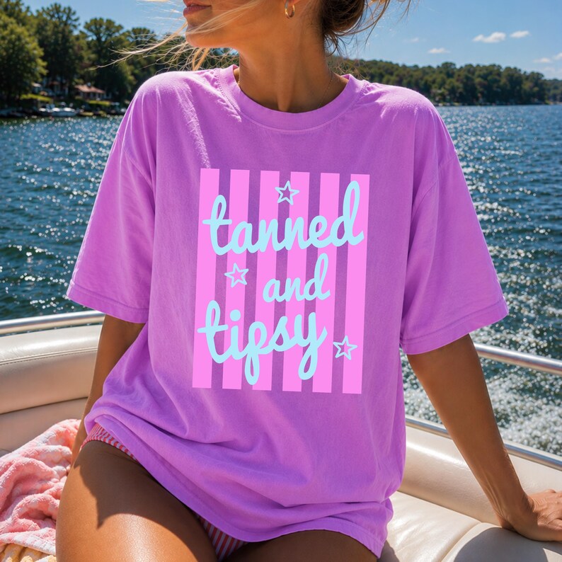 May include: A lilac-colored t-shirt with the words "tanned and tipsy" in light blue script, set against a pink striped background. The shirt has a relaxed fit and short sleeves. The image is taken outdoors with a body of water in the background.