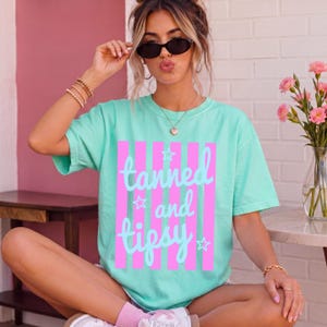 May include: A mint green t-shirt with the words "tanned and tipsy" in light blue script and stars, set against pink vertical stripes. The shirt is a casual, oversized style.