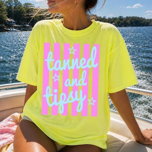 May include: A yellow t-shirt with the words "tanned and tipsy" in light blue script, set against pink vertical stripes. The shirt has a relaxed fit and short sleeves, perfect for a sunny day.