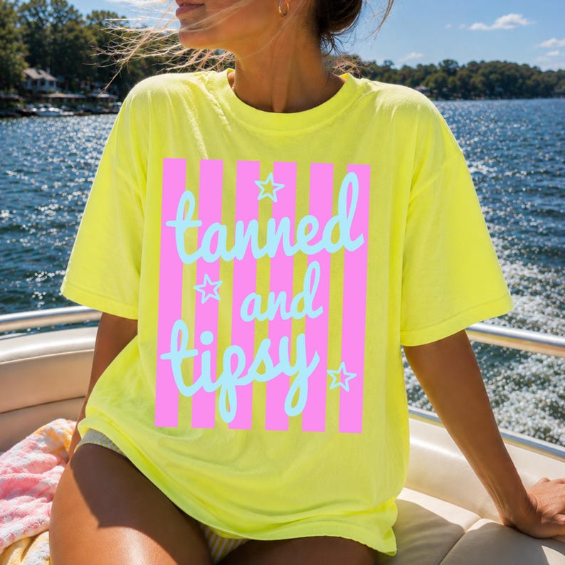 May include: A yellow t-shirt with the words "tanned and tipsy" in light blue script, set against pink vertical stripes. The shirt has a relaxed fit and short sleeves, perfect for a sunny day.