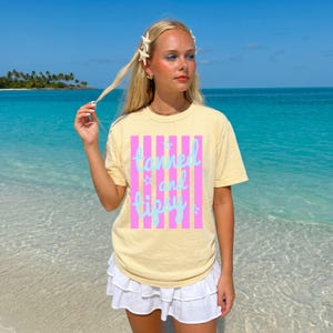 May include: A light yellow t-shirt with the words "tanned and tipsy" in a vertical striped design of pink and light blue. The shirt is worn with a white skirt, and the background is a tropical beach scene.