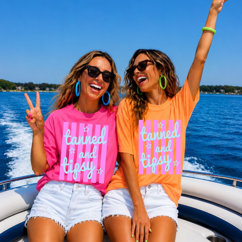 May include: Two people wearing sunglasses and matching t-shirts with the text "tanned and tipsy" in pink and blue. One shirt is pink, the other is orange. They are on a boat on a sunny day.