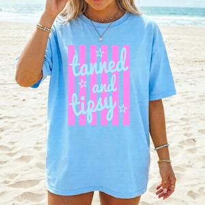 May include: Light blue t-shirt with the text "tanned and tipsy" in a cursive font, framed by pink vertical stripes. The shirt has a relaxed fit and is suitable for casual wear. The design includes star accents.