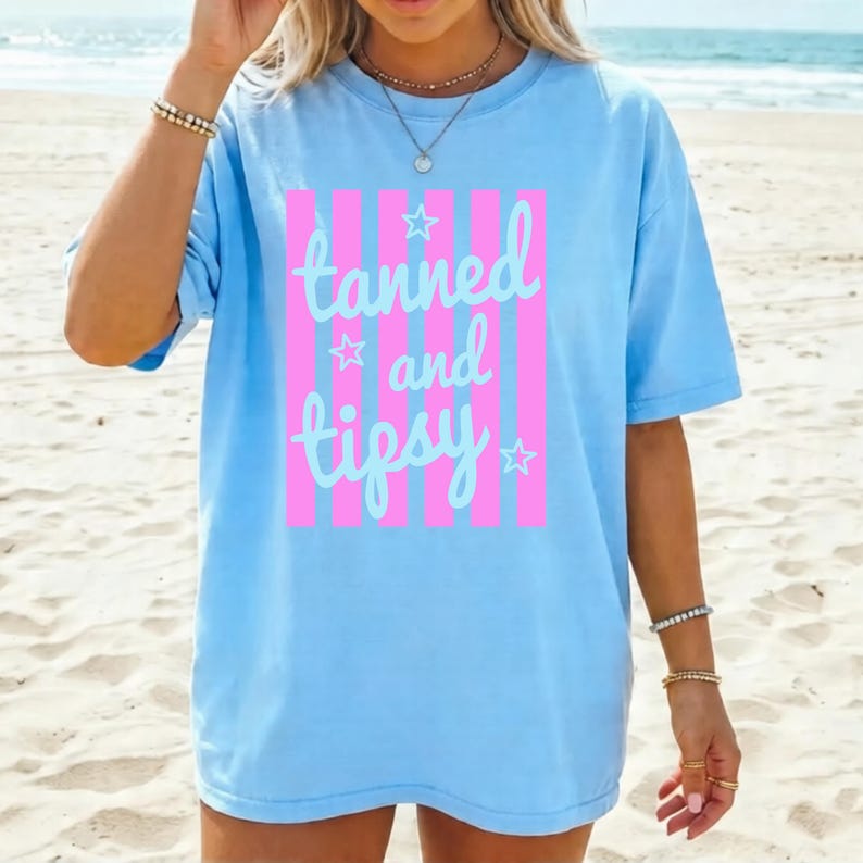 May include: Light blue t-shirt with the text "tanned and tipsy" in a cursive font, framed by pink vertical stripes. The shirt has a relaxed fit and is suitable for casual wear. The design includes star accents.