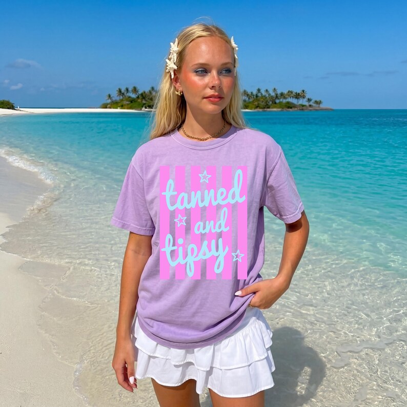May include: A lilac-colored t-shirt with the words "tanned and tipsy" in a light blue script, set against a pink striped background. The shirt is paired with a white ruffled skirt. The scene is set on a beach with turquoise water.