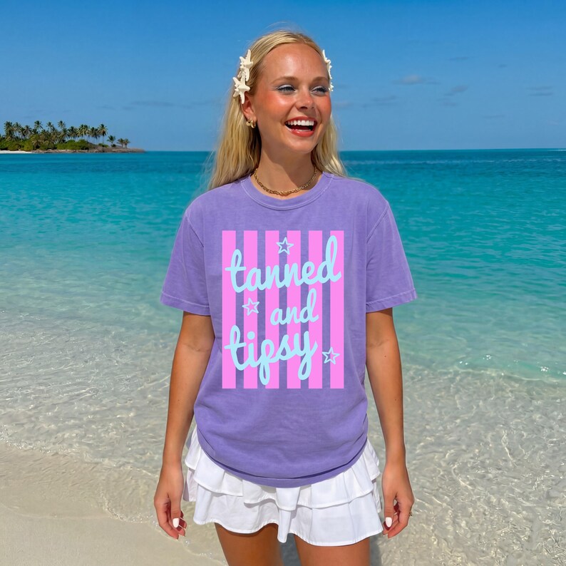May include: A purple t-shirt with the text "tanned and tipsy" in a light blue script, set against pink vertical stripes. The shirt is worn with a white skirt, against a backdrop of a tropical beach with turquoise water and blue sky.