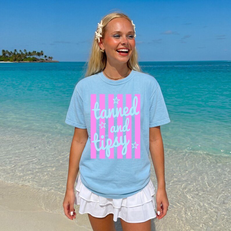 May include: A light blue t-shirt with the words "tanned and tipsy" in a pink and white striped design. The shirt is worn by a person on a beach with blue water and a blue sky.