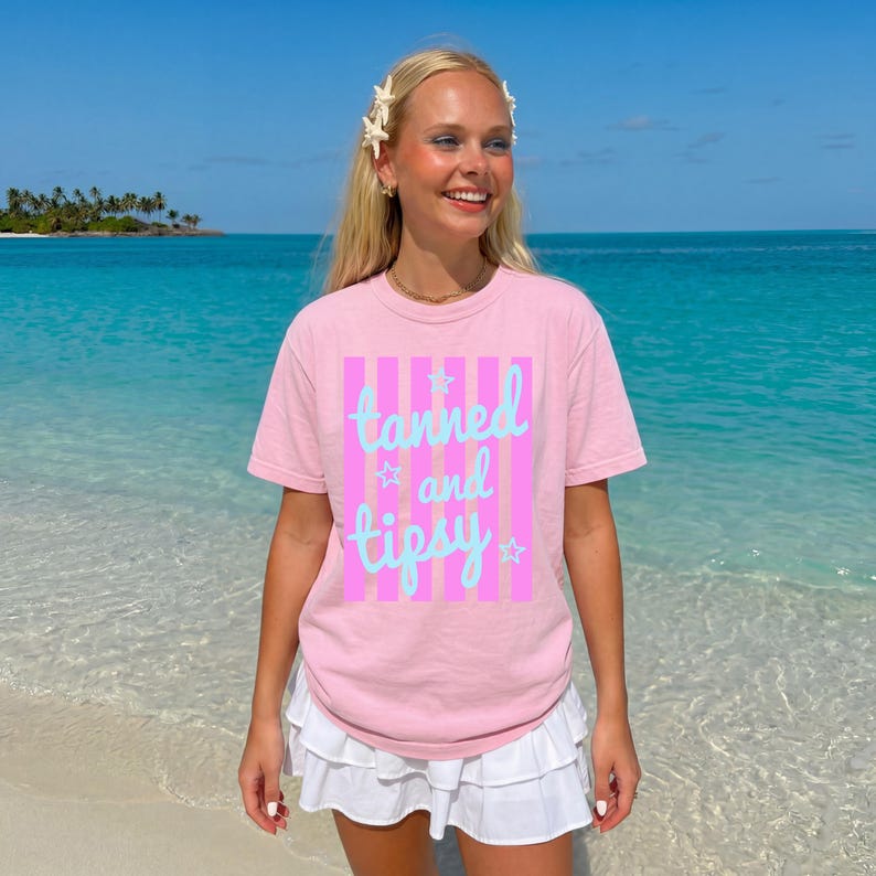 May include: A light pink t-shirt with the words "tanned and tipsy" in a light blue script, set against a pink and white striped background. The shirt is worn with a white skirt. The background shows a beach and ocean.