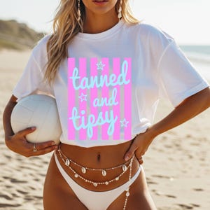 May include: White t-shirt with the text "tanned and tipsy" in light blue script over a pink striped background. The shirt is paired with a white bikini bottom and a seashell body chain. A white volleyball is held in the hand.