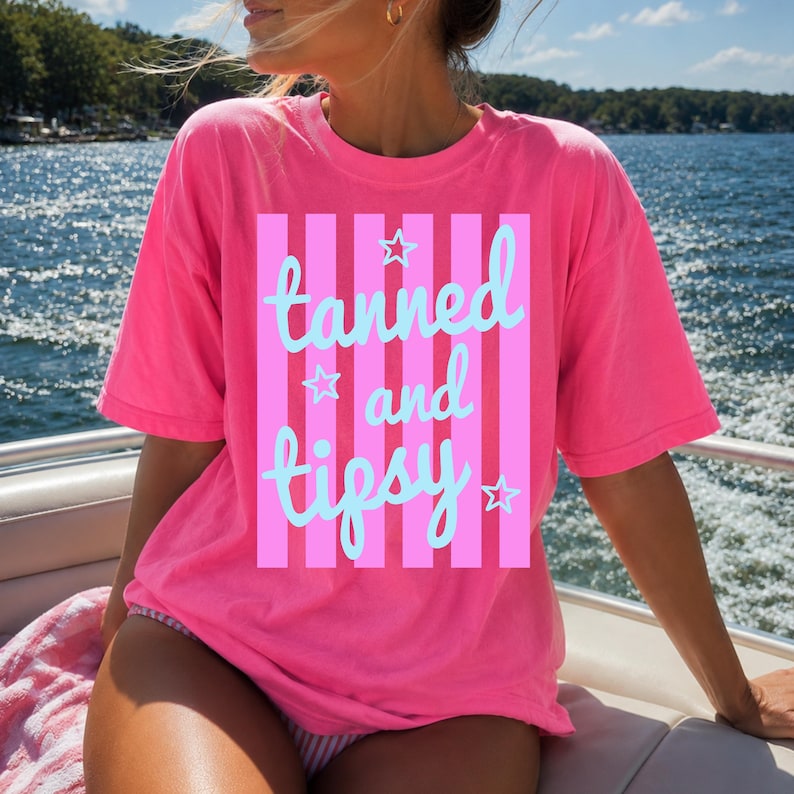 May include: A bright pink t-shirt with the words "tanned and tipsy" in light blue script and stars, set against a pink and light pink striped background. The shirt is short-sleeved.