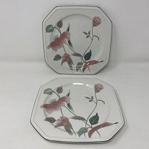 May include: Two octagonal white plates with a floral design. The plates feature pink flowers, green leaves, and gray stems. The edges are outlined with a dark blue trim. A classic dinnerware set.