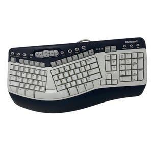 Puede incluir: Teclado ergonómico Microsoft con diseño dividido. El teclado presenta una combinación de colores negro y plateado con teclas blancas. Tiene una forma curva e incluye botones de control multimedia.