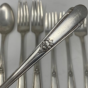 Può includere: Primo piano di posate antiche, tra cui forchette e un cucchiaio. Le posate sono di colore argento con un motivo floreale sul manico del cucchiaio. Le forchette hanno un design semplice ed elegante. Le posate sono disposte su una superficie bianca.