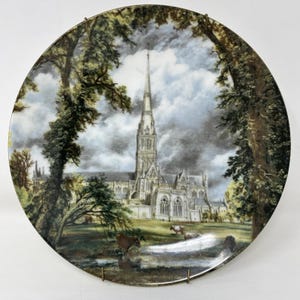 May include: A decorative plate featuring a detailed landscape painting of a cathedral with a tall spire, set amidst trees and a cloudy sky. The plate has a gold rim and is mounted on a wire stand, showcasing a pastoral scene with cows grazing near a stream.