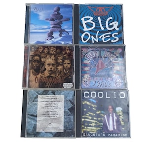 May include: A collection of six music CDs. The album covers feature diverse artwork, including a stone sculpture, a band logo, and portraits. Titles include "Big Ones," "Gangsta's Paradise," and "Hard Rock Essentials 1980s."