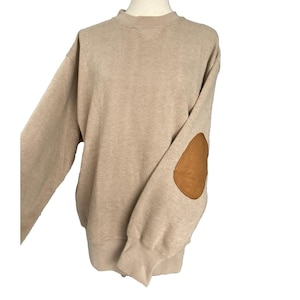 May include: Beige long-sleeve sweatshirt with a crew neck and brown elbow patches. The sweatshirt has a ribbed texture and a relaxed fit. The sleeves have ribbed cuffs. The elbow patches are oval-shaped.