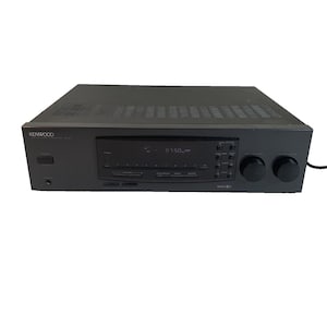 Pode incluir: Um receptor estéreo Kenwood preto com um visor digital mostrando "87.50". O receptor tem vários botões e botões, e o logotipo da Kenwood é visível no lado esquerdo.