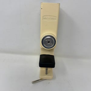 May include: A cream-colored clamp with a black pad and a silver metal screw. The text "BACK TO BASICS" is printed on the top section of the clamp. The clamp is designed to hold objects.