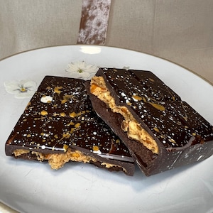 Könnte beinhalten: Dunkle Schokoladenriegel mit Erdnussbutterfüllung, beträufelt mit weißen und goldenen Akzenten, werden auf einem weißen Teller präsentiert. Die Schokolade ist angeschnitten, um die Füllung zu zeigen. Das Bild zeigt einen Gourmet-Leckerbissen.