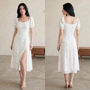 May include: A white midi dress featuring short puff sleeves, a sweetheart neckline, and a high front slit. The fabric is adorned with a delicate, scattered pattern of small red and green floral motifs, with a fitted bodice and a flowing skirt.