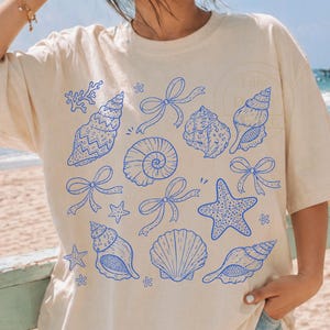 Op de afbeelding: Crèmekleurig T-shirt met een blauw schelp- en strikpatroon. Het shirt met korte mouwen heeft een relaxte pasvorm. Het ontwerp bevat verschillende schelpenillustraties en strikaccenten, wat een strandthema creëert.