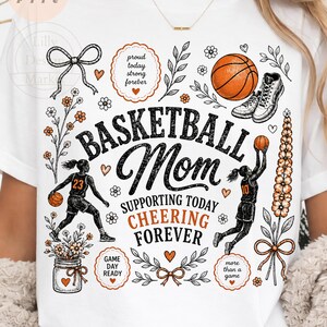 Puede incluir: Camiseta blanca con un dise&ntilde;o tem&aacute;tico de baloncesto. El dise&ntilde;o incluye las palabras "Basketball Mom" con texto de apoyo, ilustraciones de baloncesto y detalles florales. La camiseta tiene cuello redondo y mangas cortas.