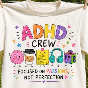 Puede incluir: Una camiseta de color crema con un gráfico colorido con el texto ADHD CREW y FOCUSED ON PASSIONS, NOT PERFECTION. El diseño incluye ilustraciones de dibujos animados de un cerebro, una taza de café, una lista de tareas amarilla, auriculares y un cubo.