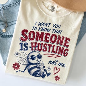 Pode incluir: Uma t-shirt cor de creme com um gráfico de uma abelha a usar óculos de sol e a segurar uma bebida. O texto diz: I WANT YOU TO KNOW THAT SOMEONE IS HUSTLING not me. O design inclui flores vermelhas e azuis e um coração.