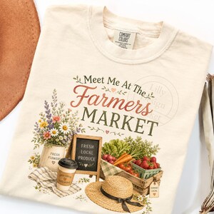 May include: Cream-colored t-shirt with a farmers market design. The design includes text that reads "Meet Me At The Farmers Market" with illustrations of flowers, produce, a chalkboard, a straw hat, and a coffee cup.