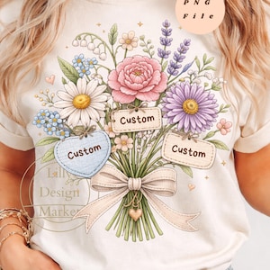 以下が含まれることがあります： クリーム色のTシャツで、カラフルな花束のイラストと大きなベージュのリボンが描かれています。デザインには「Custom」という文字が印刷された3つのラベルと、小さなハートや星のモチーフが含まれています。