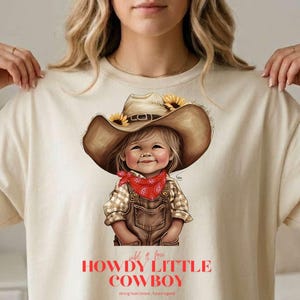 May include: A beige t-shirt featuring a graphic of a smiling child dressed as a cowboy. The child wears a brown cowboy hat with sunflowers, a red bandana, and brown overalls. The text "HOWDY LITTLE COWBOY" is printed below the image.