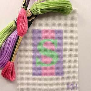 May include: A needlepoint canvas featuring a colorful letter "S" in green, pink, and purple. The canvas is accompanied by skeins of embroidery floss in pink, green, and purple. The craft supplies are ideal for a DIY project.