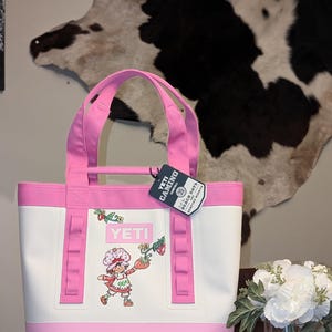 May include: A white and pink YETI Camino Carryall bag with pink handles. The bag features a Strawberry Shortcake graphic and the YETI logo. The bag has pink accents and a tag that reads "YETI CAMINO".