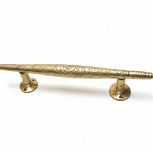 May include: A brass door pull with an ornate, engraved floral design. The handle is a warm gold color and has a cylindrical shape with decorative detailing. The pull has two circular mounting plates on each end.