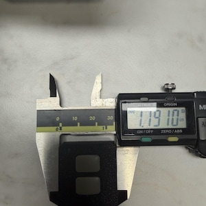 May include: A black rectangular object with two recessed buttons is measured with a digital caliper. The caliper displays a measurement of 1.1910 inches. The caliper is silver and black, and the object is on a light gray surface.