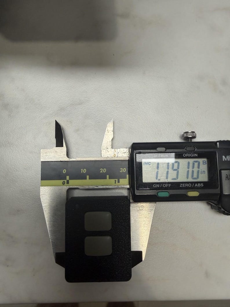 May include: A black rectangular object with two recessed buttons is measured with a digital caliper. The caliper displays a measurement of 1.1910 inches. The caliper is silver and black, and the object is on a light gray surface.