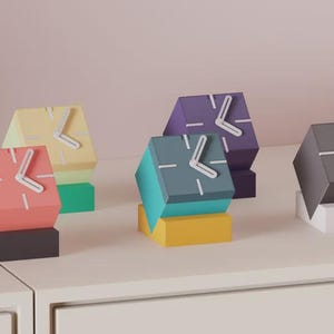 May include: A collection of colorful, geometric cube-shaped clocks. Each clock features a different color, including pink, yellow, teal, purple, and gray. The clocks have white hands and are displayed on a white surface.