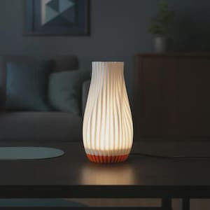 May include: A white, ribbed table lamp with an orange base, illuminated against a dark background. The lamp has a vase-like shape and is plugged into an electrical outlet. The lamp is on a dark table.