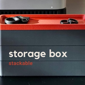 May include: A stackable storage box with a red top and black body. The words "storage box" are printed in white, and "stackable" is in red. Two black rings and a black clip rest on the top.