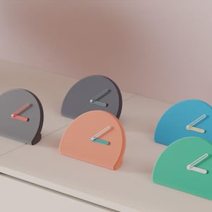 Peut inclure: Plusieurs horloges de bureau modernes, semi-circulaires, de couleurs variées : gris, pêche, bleu et vert. Chaque horloge a des aiguilles de couleurs contrastées. Les horloges sont disposées sur une surface blanche, sur fond rose pâle.