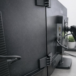 Könnte beinhalten: Schwarze Computermonitore mit angebrachten schwarzen Halterungen. Die Halterungen sind an der Rückseite der Monitore befestigt und scheinen aus Kunststoff zu sein. Kabel sind an die Monitore angeschlossen.