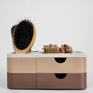 Puede incluir: Organizador de varios compartimentos en tonos marrón, beige y blanco. Cuenta con dos cajones y una sección superior que contiene un cepillo para el cabello con mango de madera, una pinza para el pelo y coleteros. El organizador tiene una textura acanalada.