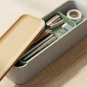 Könnte beinhalten: Ein rechteckiger, geriffelter, hellgrauer Schreibtisch-Organizer mit beigem Deckel. Im Inneren befindet sich eine grüne Ablage mit Tacker, Bleistiften, Stiften, Büroklammern, Radiergummi und Klebeband. Der Deckel ist teilweise geöffnet.